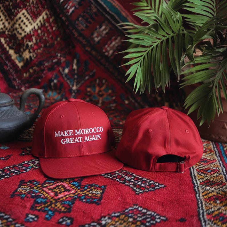 Op de afbeelding: Twee rode baseballpetten. Een pet heeft de tekst "MAKE MOROCCO GREAT AGAIN" in wit geborduurd op de voorkant.