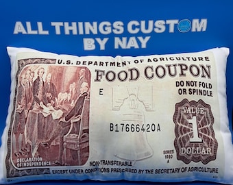 Może przedstawiać: Biała poduszka z nadrukowanym na niej bonem żywnościowym w stylu vintage. Bon jest wart jednego dolara i zawiera tekst "U.S. Department of Agriculture Food Coupon" oraz "B17666420A".