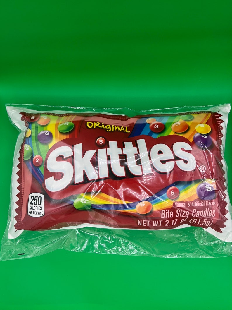 Op de afbeelding: Een rode, witte en regenboogkleurige zak Skittles Original Bite Size Candies. De zak heeft een wit label met het Skittles-logo en de tekst "Original" en "Natuurlijke en kunstmatige smaken". De zak heeft ook een voedingswaardelabel met de tekst "250 calorie&euml;n per portie".