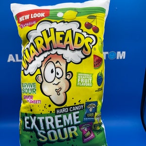 Puede incluir: Una almohada blanca con forma de bolsa de caramelos duros Warheads Extreme Sour. La bolsa es amarilla y verde con un personaje de dibujos animados en la parte delantera. El texto en la bolsa dice "New Look Extreme Warheads Hard Candy Extreme Sour" y "Peso neto 2 oz (56 g)".