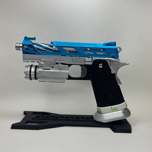 May include: A silver and blue pistol with a black grip and a green sight. The pistol is on a black stand and has the text "3011 TYPE 5015" on the side.