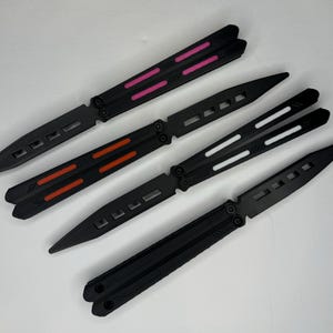 May include: Three black butterfly knives with different colored accents. The first knife has pink accents, the second has orange accents, and the third has white accents.