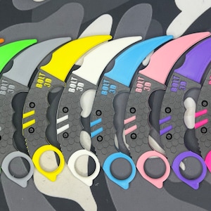 Pode incluir: Um conjunto de 10 facas karambit com alças de diferentes cores. As alças são de plástico e as lâminas de metal. As facas estão todas em fila e as lâminas estão orientadas na mesma direção. As facas são todas do mesmo tamanho e forma. As facas são todas pretas com um padrão de favo de mel na lâmina. As alças são de diferentes cores, incluindo azul, laranja, verde, cinza, amarelo, branco, azul claro, rosa, roxo e vermelho.