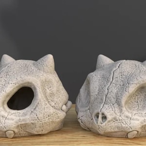 Puede incluir: Dos objetos decorativos con forma de calavera blanquecina, con grandes cuencas oculares y superficies texturizadas. Las calaveras tienen orejas puntiagudas y están colocadas sobre una superficie de madera contra un fondo gris oscuro. Los objetos parecen estar hechos de un material similar a la piedra.