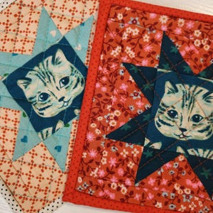 May include: Two quilted fabric squares. One features a cat face in a blue star on a light blue background with hearts. The other has a cat face in a blue star on a floral background. Both have red borders.