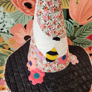 May include: A quilted cone-shaped hat featuring a floral pattern with pink, orange, and green colors. The hat displays a large appliqued bee with white wings and a yellow and black striped body. The wide brim is black with a white spiderweb pattern.