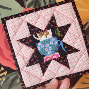 May include: A square quilted potholder featuring a star design. The star is black with a blue teacup illustration and pink floral accents. The background is white with pink polka dots, and the edges are black with pink floral details.