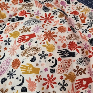 May include: White fabric with a repeating pattern of colorful moths, flowers, hands with hearts, stars, and crescent moons. The design features shades of orange, red, pink, yellow, black, and navy blue.