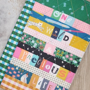 Könnte beinhalten: Ein gestepptes Stoffstück mit einem Patchwork-Design. Das Design umfasst verschiedene Muster, wie Punkte, Blumenmuster und Vichy-Karos. Die Worte "ONE WILD & PRECIOUS LIFE" sind in das Design integriert. Der Artikel ist mit einem Rautenmuster genäht.