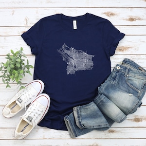 Puede incluir: Una camiseta azul marino con un contorno blanco de un mapa de la ciudad. La camiseta está colocada sobre una superficie de madera blanca con un par de zapatillas blancas y unos vaqueros azules.