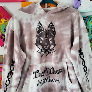 Op de afbeelding: Een wit en bruin tie-dye hoodie met een zwarte afbeelding van een hondenkop met een stekelband. De voorzak heeft de tekst "Them Mayhem", en de mouwen hebben een kettingontwerp. Een casual kledingstuk.