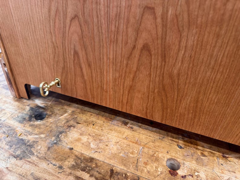 May include: Close-up of a wooden cabinet with a keyhole and a decorative brass key. The cabinet is made of reddish-brown wood with visible grain patterns. The key is attached to the cabinet's side.