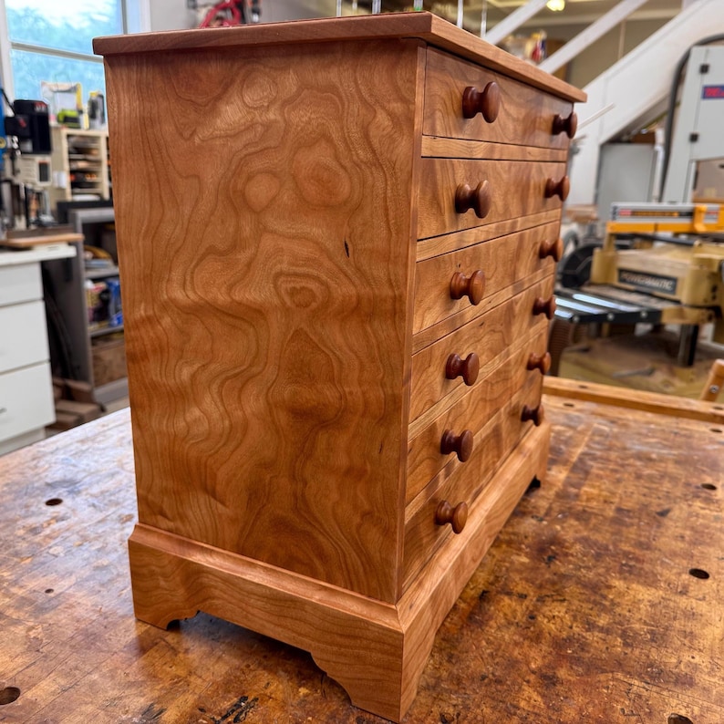 May include: A small, wooden chest of drawers with five drawers and round wooden knobs. The chest is made of reddish-brown wood with a visible wood grain pattern. The chest sits on a base with decorative feet.