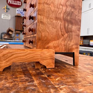 May include: A handcrafted wooden cabinet with multiple drawers and a matching tray. The cabinet is made of reddish-brown wood with a visible grain pattern. The drawers have dark brown knobs. The tray is placed in front of the cabinet.