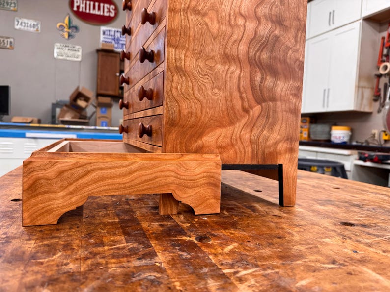 May include: A handcrafted wooden cabinet with multiple drawers and a matching tray. The cabinet is made of reddish-brown wood with a visible grain pattern. The drawers have dark brown knobs. The tray is placed in front of the cabinet.