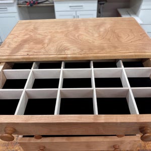 May include: Wooden jewelry box with multiple compartments. The box is made of light-colored wood with a natural finish. The interior features a black velvet lining and white dividers. The box has two drawers with round wooden knobs.