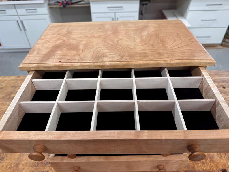 May include: Wooden jewelry box with multiple compartments. The box is made of light-colored wood with a natural finish. The interior features a black velvet lining and white dividers. The box has two drawers with round wooden knobs.