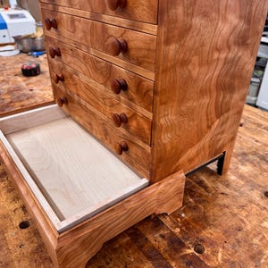 May include: A small wooden chest of drawers with a warm, reddish-brown finish. The chest has four drawers with round wooden knobs and a pull-out tray at the bottom. The wood grain is visible, and the chest sits on small black legs.