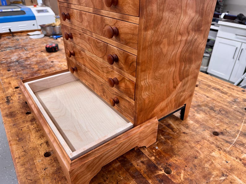 May include: A small wooden chest of drawers with a warm, reddish-brown finish. The chest has four drawers with round wooden knobs and a pull-out tray at the bottom. The wood grain is visible, and the chest sits on small black legs.