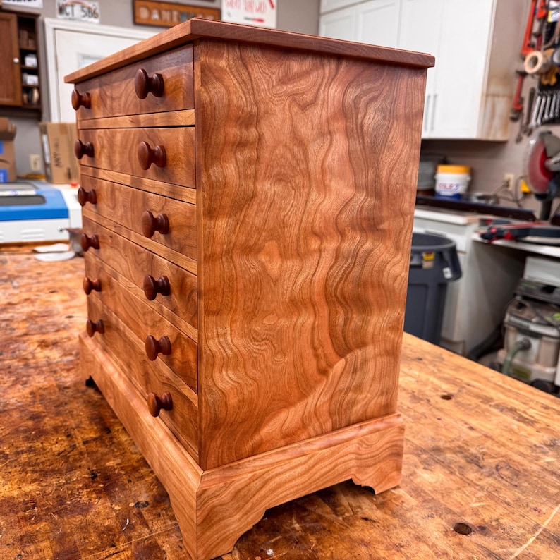 May include: A handcrafted wooden chest of drawers with a warm, reddish-brown finish. The chest features five drawers with rounded wooden knobs. The wood grain is prominently displayed, adding to the piece's natural aesthetic. The chest sits on a decorative base.