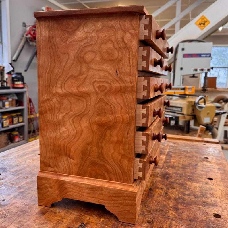 May include: A small, wooden chest of drawers with five drawers. The chest is made of reddish-brown wood with a visible wood grain pattern. Each drawer has a small, round wooden knob. The chest sits on a base with decorative feet.