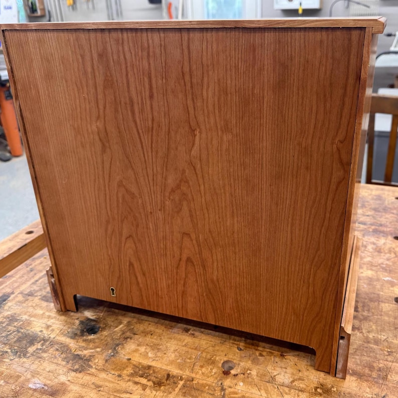May include: A wooden cabinet with a reddish-brown finish. The cabinet has a rectangular shape with visible wood grain patterns. The back of the cabinet is shown, with a small keyhole near the bottom. The cabinet is sitting on a wooden surface.
