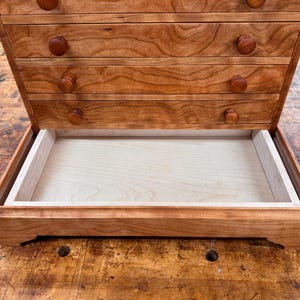 May include: A wooden jewelry box with a cherry finish. The box has multiple drawers with round wooden knobs and a large open tray at the bottom. The wood grain is visible, and the box sits on a wooden surface.