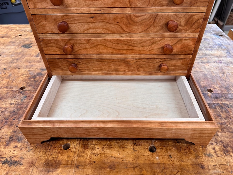 May include: A wooden jewelry box with a cherry finish. The box has multiple drawers with round wooden knobs and a large open tray at the bottom. The wood grain is visible, and the box sits on a wooden surface.