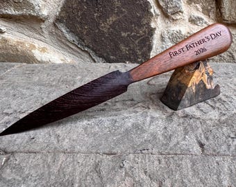 May include: A handcrafted letter opener with a dark blade and a wooden handle engraved with "FIRST FATHER'S DAY 2026." The opener rests on a small, decorative wooden stand, showcasing a blend of dark and light wood tones.