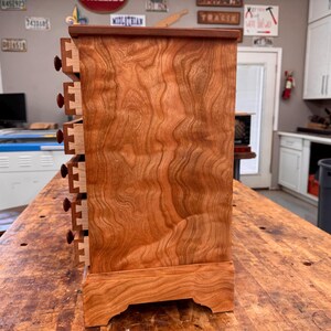 May include: A handcrafted wooden cabinet with six small drawers. The cabinet is made of reddish-brown wood with a visible wood grain pattern. Each drawer has a small, round, dark brown knob. The cabinet sits on a decorative base.