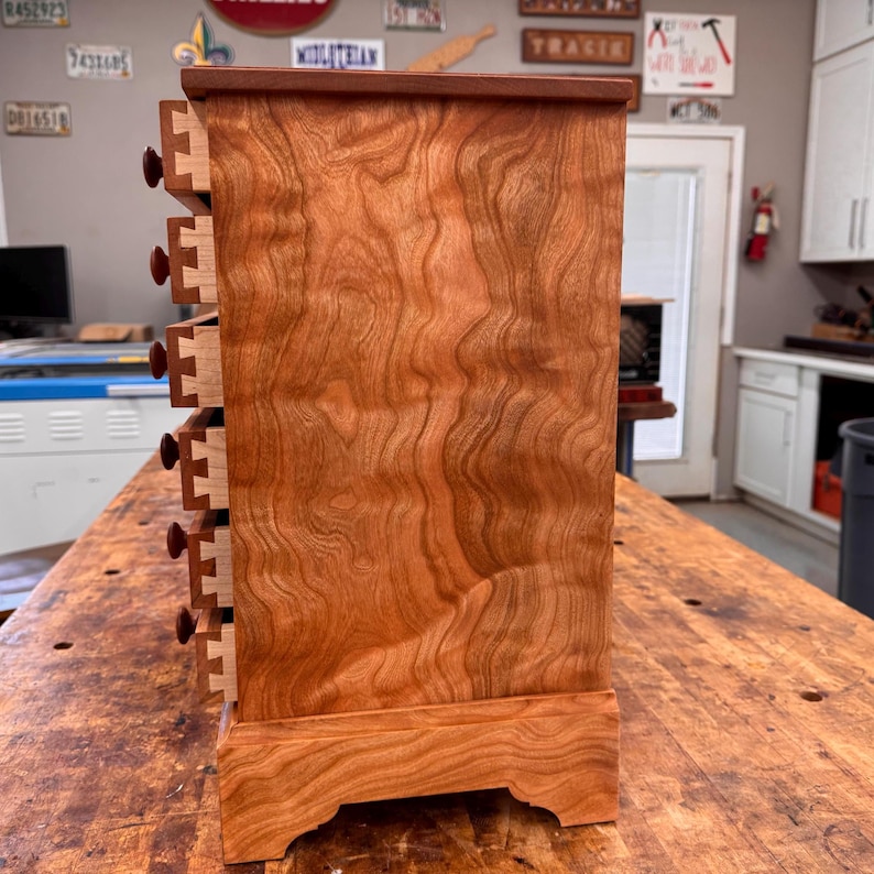 May include: A handcrafted wooden cabinet with six small drawers. The cabinet is made of reddish-brown wood with a visible wood grain pattern. Each drawer has a small, round, dark brown knob. The cabinet sits on a decorative base.