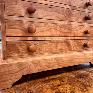 May include: A small, wooden chest of drawers with five drawers. The chest is made of reddish-brown wood with a natural grain pattern. Each drawer has a round, wooden knob. The chest sits on a decorative base.