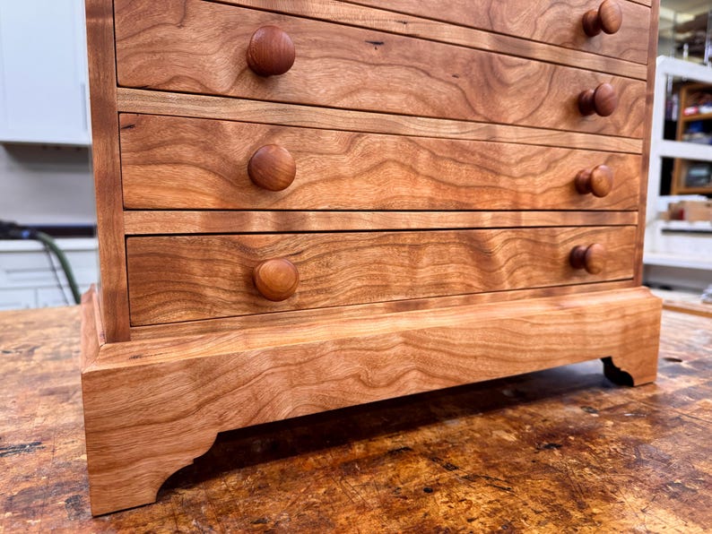 May include: A small, wooden chest of drawers with five drawers. The chest is made of reddish-brown wood with a natural grain pattern. Each drawer has a round, wooden knob. The chest sits on a decorative base.