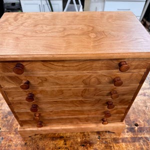 May include: A wooden chest of drawers with five drawers and round wooden knobs. The chest is made of light brown wood with a natural grain pattern. The chest sits on a small base and has a flat top.