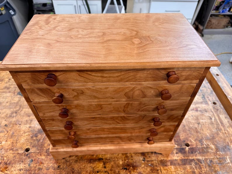 May include: A wooden chest of drawers with five drawers and round wooden knobs. The chest is made of light brown wood with a natural grain pattern. The chest sits on a small base and has a flat top.