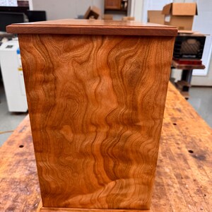 May include: A handcrafted wooden box with a rich, reddish-brown finish. The box features a rectangular shape with a slightly tapered design and a decorative base. The wood grain is prominent, adding to the natural aesthetic. The box is likely used for storage or display.