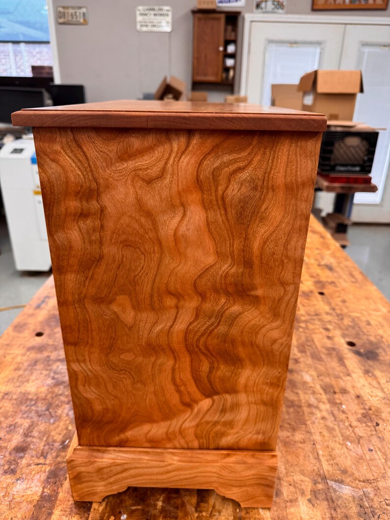 May include: A handcrafted wooden box with a rich, reddish-brown finish. The box features a rectangular shape with a slightly tapered design and a decorative base. The wood grain is prominent, adding to the natural aesthetic. The box is likely used for storage or display.