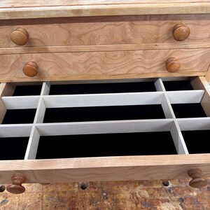 May include: A wooden jewelry box with two drawers and a pull-out compartment. The box is made of light-colored wood with a natural finish. The drawers have round wooden knobs. The open compartment has black velvet lining and white dividers.
