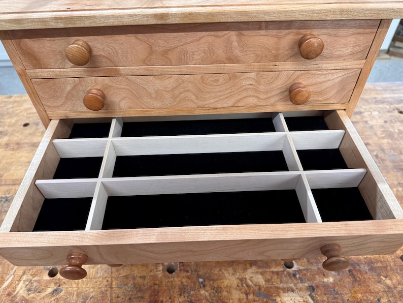 May include: A wooden jewelry box with two drawers and a pull-out compartment. The box is made of light-colored wood with a natural finish. The drawers have round wooden knobs. The open compartment has black velvet lining and white dividers.