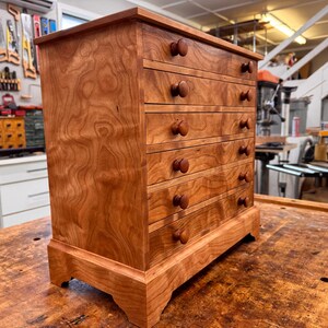May include: A small, wooden chest of drawers with five drawers and round wooden knobs. The chest is made of reddish-brown wood with a visible wood grain pattern. It sits on a wooden surface, showcasing the craftsmanship of the piece.