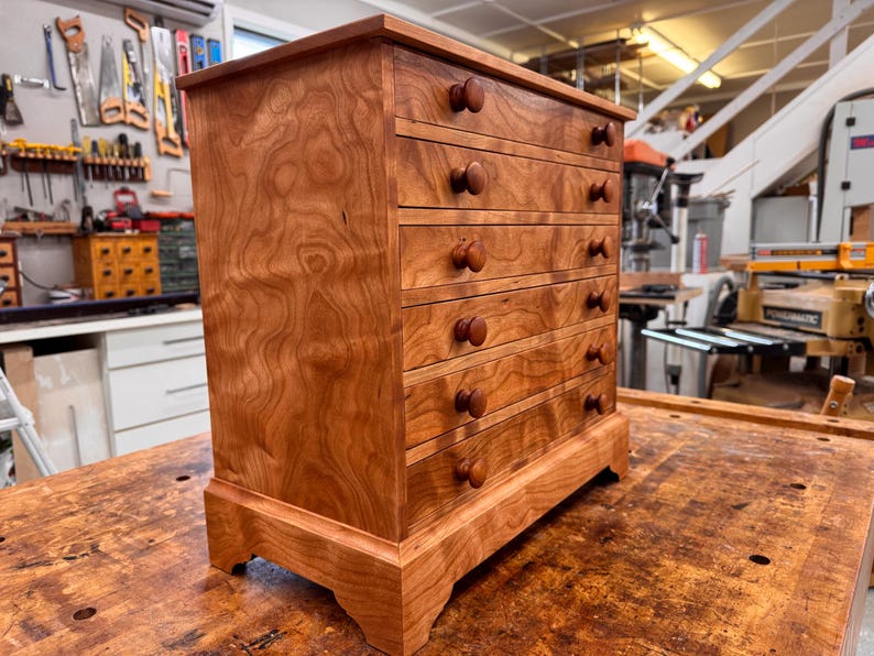May include: A small, wooden chest of drawers with five drawers and round wooden knobs. The chest is made of reddish-brown wood with a visible wood grain pattern. It sits on a wooden surface, showcasing the craftsmanship of the piece.