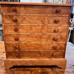 May include: A small, wooden chest of drawers with six drawers. The chest is made of light brown wood with a visible wood grain. Each drawer has a round, wooden knob. The chest sits on a small base.