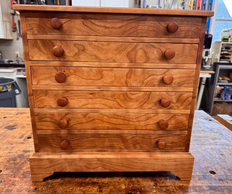 May include: A small, wooden chest of drawers with six drawers. The chest is made of light brown wood with a visible wood grain. Each drawer has a round, wooden knob. The chest sits on a small base.