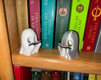 May include: Two ghost-shaped bookends, one white and one silver, each holding a small blue book. They sit on a wooden shelf in front of a row of colorful books. A unique and whimsical addition to any bookshelf.