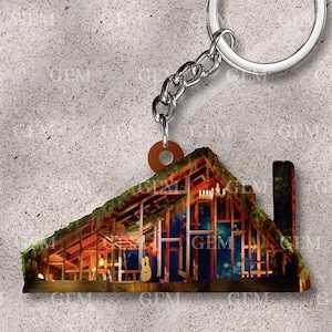 Op de afbeelding: Een houten sleutelhanger met een gedetailleerd beeld van een gebouw met een groen dak en een gitaar erin. De sleutelhanger is bevestigd aan een zilveren metalen ring en ketting.