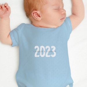 May include: A light blue baby onesie with white text that reads "2023 CHARLIE'S VERSION".