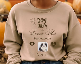May include: A woman wearing a tan sweatshirt with the text "Just a Dog Mom Who Loves Her Bernedoodle" and a photo of a dog named Nemo. The sweatshirt is in front of a truck bed with pumpkins.
