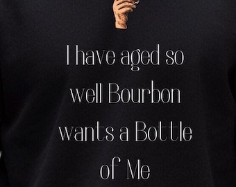 May include: Black sweatshirt with white text that reads "I have aged so well Bourbon wants a bottle of me". A small image of a hand holding a glass of bourbon is above the text.