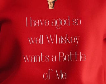 May include: Red sweatshirt with white text that reads "I have aged so well Whiskey wants a Bottle of Me". A small illustration of a hand holding a glass of whiskey is above the text.