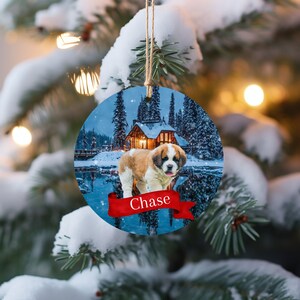 May include: Round Christmas ornament featuring a Saint Bernard puppy in a snowy landscape with a cabin and lake. The puppy has brown and white fur. A red banner at the bottom reads "Chase". The ornament hangs from a natural twine string.